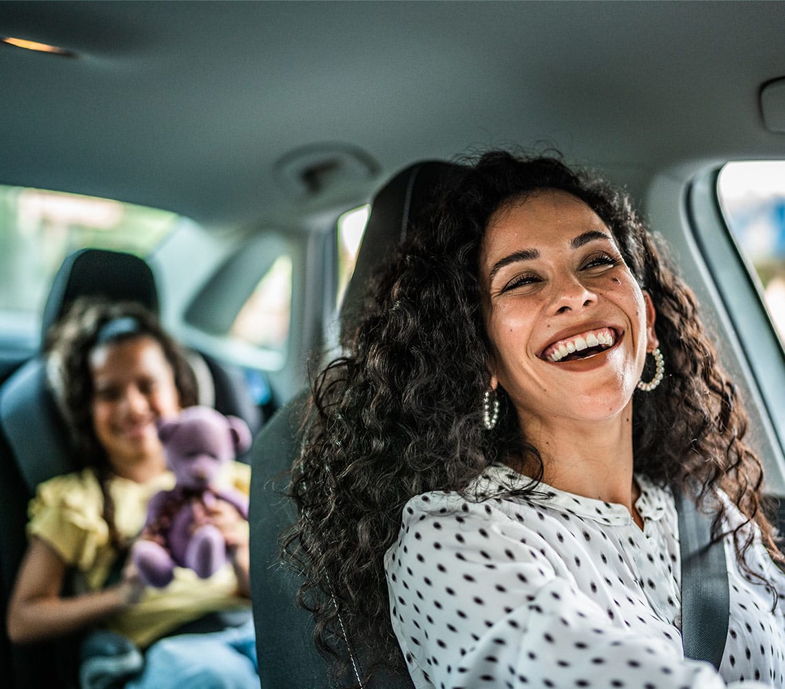 A woman driving a vehicle and smiling at her child holding a purple teddy bear sitting in the back of a vehicle.